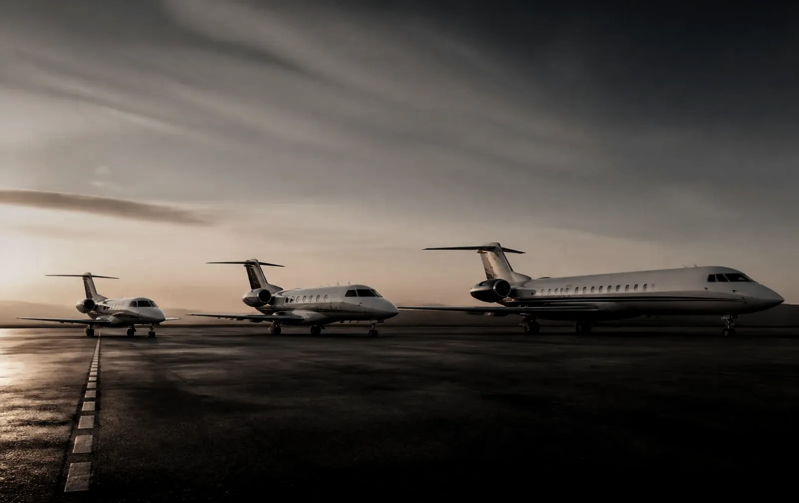 Private jets lined up on a wet airport ramp at dusk under a storm-grey sky